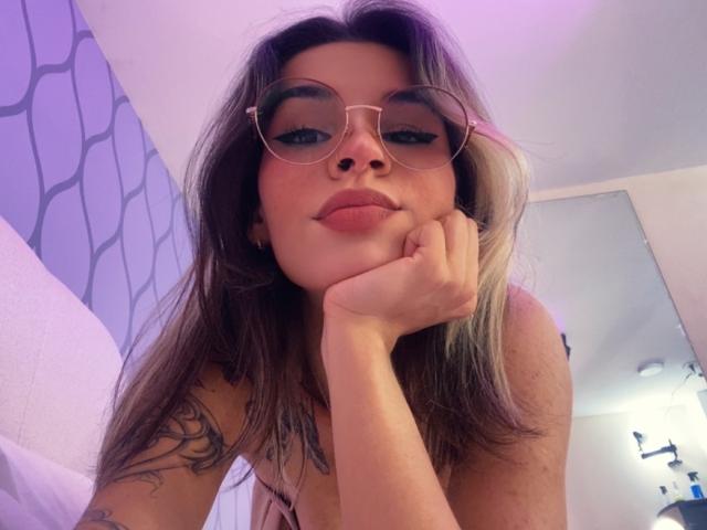 LenaBranch, con le labbra tumefatte, gli occhiali e un tatuaggio sulla spalla, appoggia il mento sulla mano, mostrando il seno e i glutei, in una camera da letto con illuminazione viola..