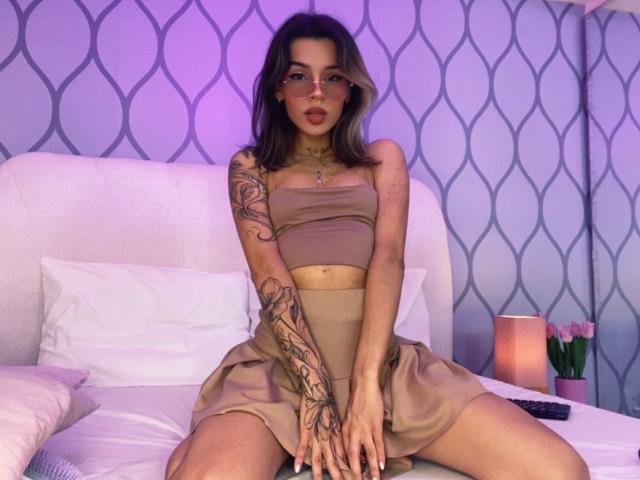 LenaBranch mit Outfit in Hauttönen, Brille und Tattoos, sitzt auf dem Bett mit gespreizten Beinen, wobei ihre Brüste und Vulva durch ihr Kleid sichtbar sind, während ihre Arme ihren Hintern bedecken. Lila Beleuchtung..