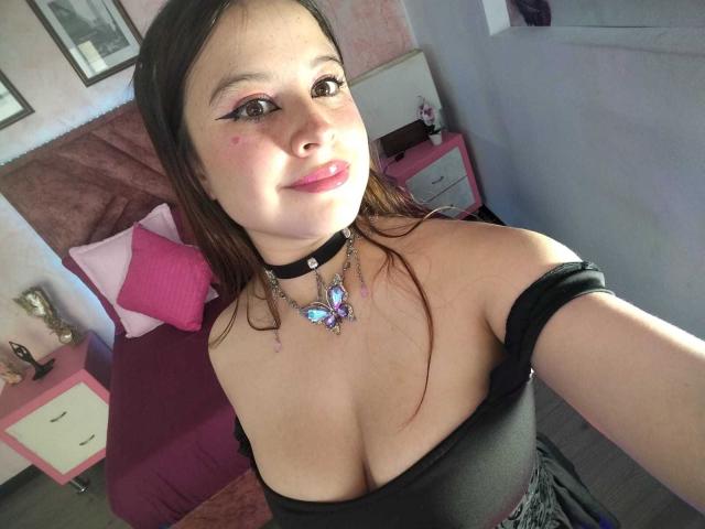 MelaniBecker with fair skin, brown eyes, and pink blush, wearing a black off-shoulder top, choker, and butterfly necklace, showing her tits in a selfie.