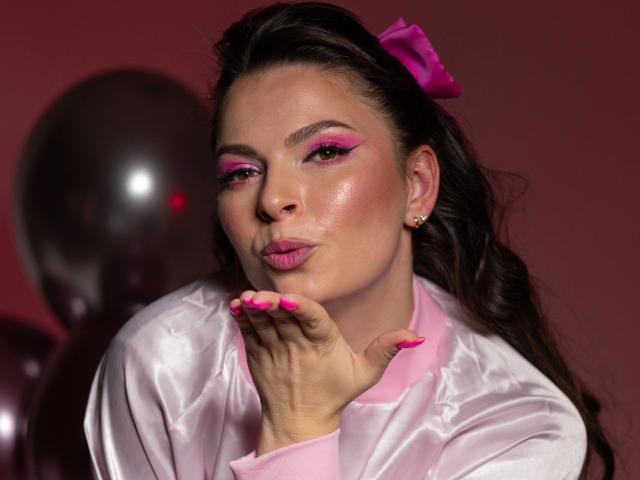 Mamasita, aantrekkelijk met roze eyeliner, glanzende roze lippen, roze nagels en een roze strik, die haar vingers kust, draagt een glanzend roze satijnjasje, met metalen ballonnen op de achtergrond..