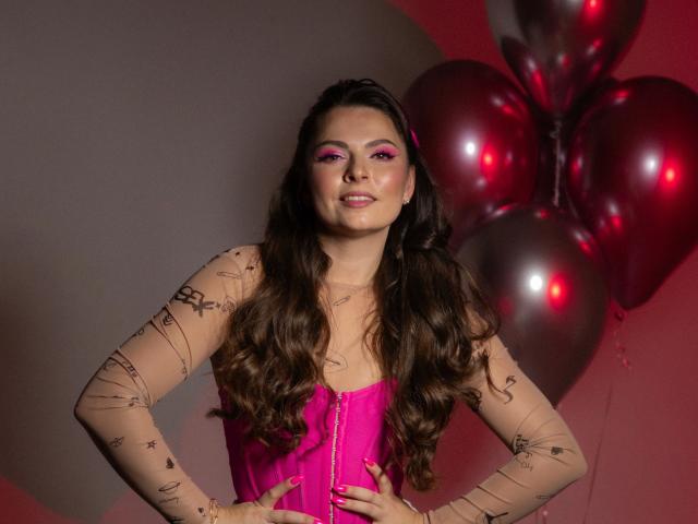 Mamasita: bella, con lunghi capelli castani, trucco rosa, indossa un corsetto rosa e maniche trasparenti con tatuaggi a linee nere, in piedi con le mani sui fianchi, palloncini argentati sullo sfondo..