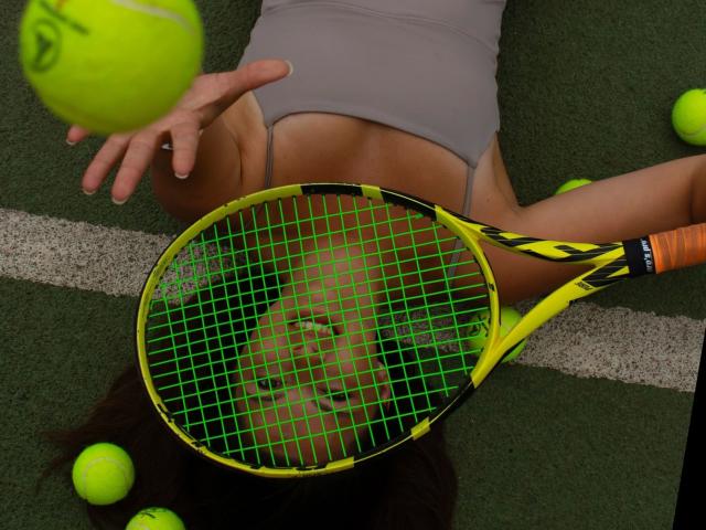 O rosto da Mamasita-hot está pressionado por uma raquete de tênis amarela, com uma bola verde neon pairando acima de sua cabeça, vestindo uma camiseta esportiva cinza, cercada por bolas de tênis..