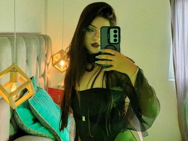 Helena Morningstar, com cabelos longos e escuros, vestindo uma blusa preta transparente, selfie no espelho, segurando um celular, mostrando os seios, travesseiro verde, luminária geométrica amarela, travesseiro azul-turquesa e cama cinza..