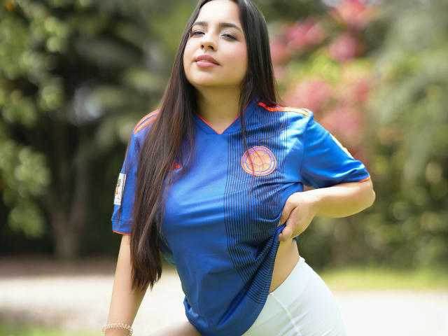 SammyTitss com cabelos longos e escuros, camisa de futebol azul, detalhes em vermelho e laranja, puxando a camisa para cima para revelar o bumbum, em pé ao ar livre, olhando para cima com confiança, luz verde suave..
