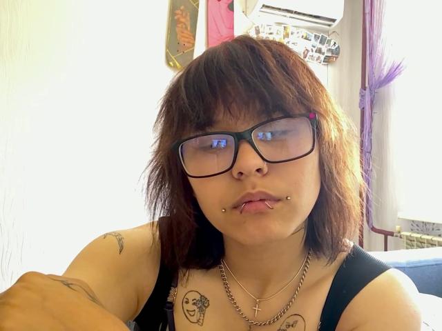 GlennisPaschel with brown hair, glasses, and nose ring, tongue piercing, tattoos, black tank top, necklace, and cross, sitting indoors.