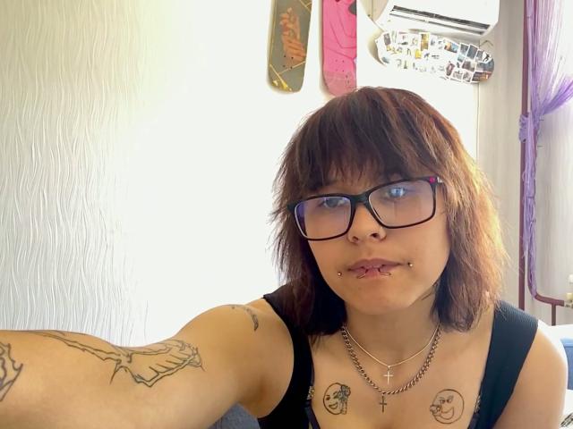 GlennisPaschel with brown hair, glasses, and nose ring, has tattoos on arms and chest, black tank top, and lip piercing, taking a selfie in a sunlit room.