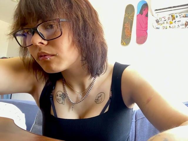 GlennisPaschel with glasses, brown shaggy hair, black tank top, tattoos on chest, nose piercing, chain necklace, sitting on couch, snowboards on wall, light-skinned, slightly messy.