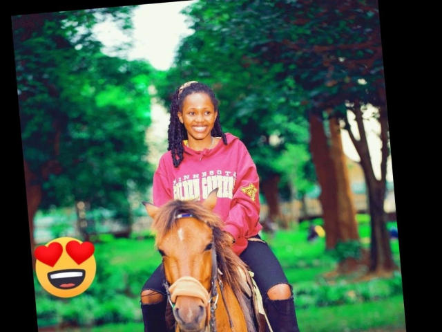 Ariyah, belle avec sa peau foncée, ses cheveux bouclés et son grand sourire, chevauche un cheval brun dans un parc verdoyant, vêtue d'un sweat à capuche rose et d'un jean noir déchiré. Émoji cœur..