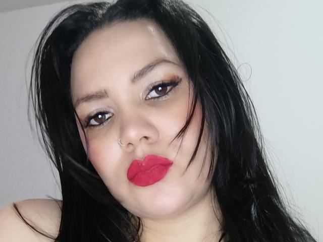 Pherlla, avec ses cheveux noirs, son rouge à lèvres rouge et son anneau de nez, a une expression légèrement boudeuse. Elle a de longs cils et un regard mignon et séduisant. Ses seins ne sont pas visibles, seulement son visage..