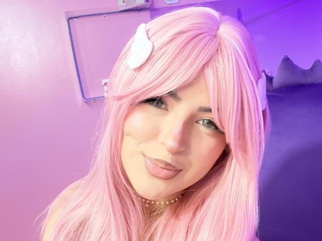 RheaVolk with pink hair, white hair clip, and pouty lips, wearing a gold choker, smiling at the camera, showing her face and upper chest.
