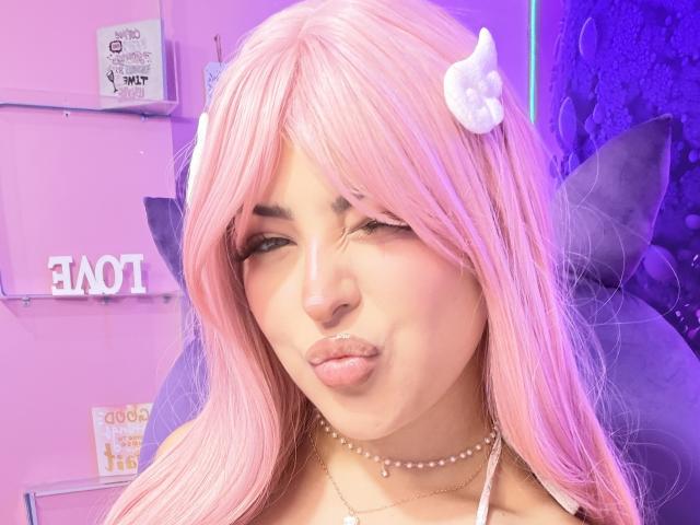 RheaVolk with pink hair, white hair clip, and pearl necklace, puckering her lips in a selfie, wearing makeup with long lashes, and a gold chain, looking cute.