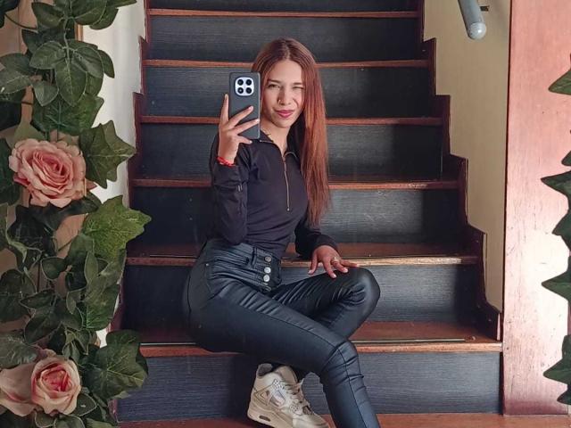 ValerieWoods, with long brown hair, wearing a black shirt and tight black leather pants, sits on stairs, holding a phone, smiling, showing her ass in white sneakers.