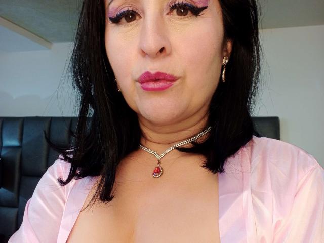 ScarletMilfX con rossetto rosa, eyeliner scuro e ombretto rosa, indossa un camice di seta rosa, una collana di diamanti e orecchini, mostrando il suo décolleté. Ha i capelli neri e un'espressione seria..