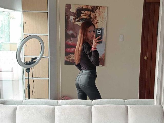 ValerieWoods, com cabelos ruivos longos, blusa preta e calças cinzas justas, aparecendo de costas em uma selfie no espelho, segurando um iPhone, com uma luz de anel à esquerda, e um sofá branco ao fundo..