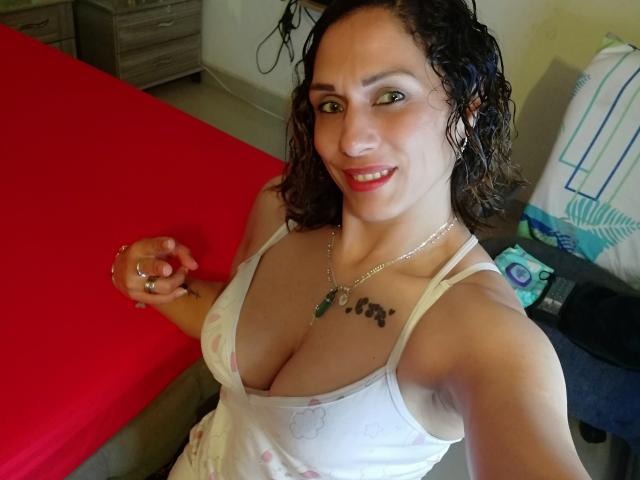 AmeliaFuentes with curly brown hair, smiling, wearing a white tank top showing her big tits, holding a red cloth, green necklace, and tattoo above her cleavage.