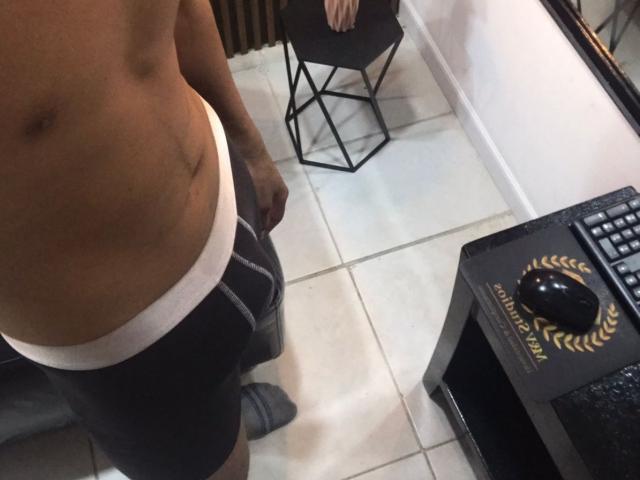 Het kale, gespierde bovenlichaam van LeleHott, met een kleine litteken, draagt zwarte boxershorts met een witte tailleband, en staat naast een zwart bureau en een stoel..