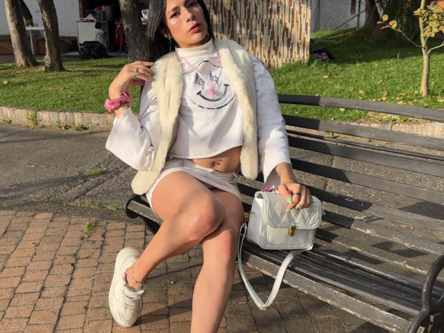 ArielNitzche with medium tits, wearing a white crop top, fur vest, and white mini-skirt, sits on a bench, legs crossed, holding a white bag, in sunlight.