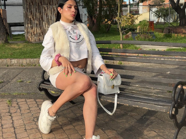 ArielNitzche with long black hair, white crop top, fluffy vest, white shorts, and sneakers, sitting on a bench, holding a white purse, showing her ass and green nails.