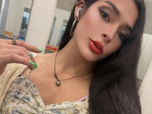 ArielNitzche with long dark hair, green nails, red lipstick, wearing a floral top, necklace, and earbuds, looking at the camera with a slight smile. Boobs visible.