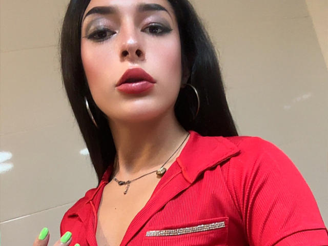 ArielNitzche with dark hair, red shirt, hoop earrings, and green nails, pouting with red lipstick, wearing a necklace, has a cute face and small tits.