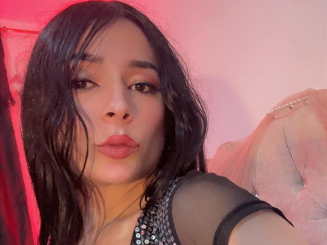 ArielNitzche has wet black hair, full red lips, and dark eyes, wearing a sheer black top with sparkly details, pouting her lips, in a pink-lit room.