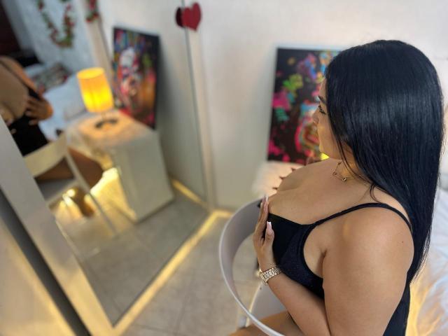 BellaaBombshell, con cabello negro largo, vestida con un sostén negro, sosteniendo sus grandes senos, mirando un espejo. Un cuadro colorido y un corazón rojo están en el fondo..