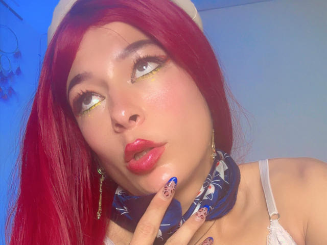 ArielNitzche with red hair, blue nail polish, gold eyeliner, and red lips, touching her lip with a finger, wearing a white hat and patterned scarf, looking up.