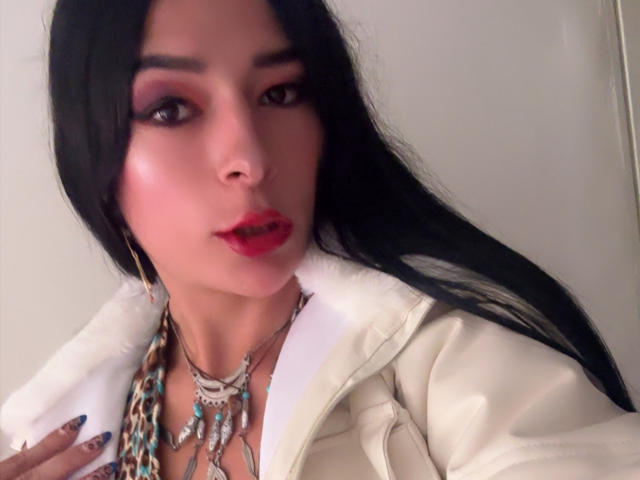 ArielNitzche with long black hair, red lipstick, and makeup, wearing a white jacket with a fur collar, and a chain necklace, looking at the camera.