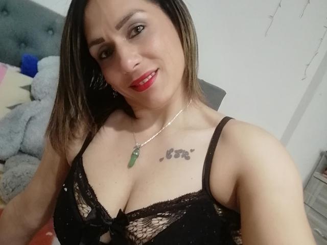 AmeliaFuentes with red lipstick, black lace bra, showing her tits, green pendant necklace, tattoo above her tits, smiling, brown hair, white wall, grey headboard, fluffy grey pillow.