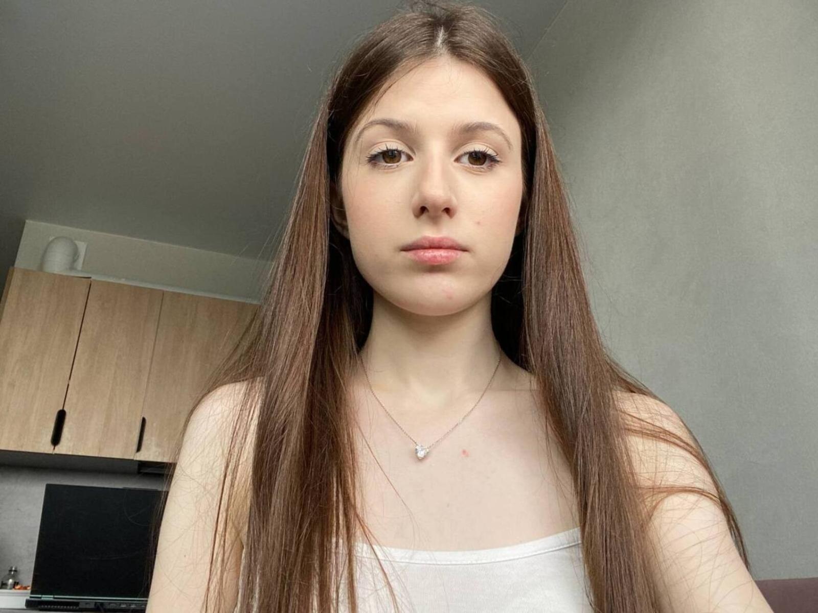 RanaeKaut heeft lang bruin haar, een lichte huid en een neutrale uitdrukking. Ze draagt een witte tanktop en een dunne ketting met een klein hangertje. Borsten zichtbaar..