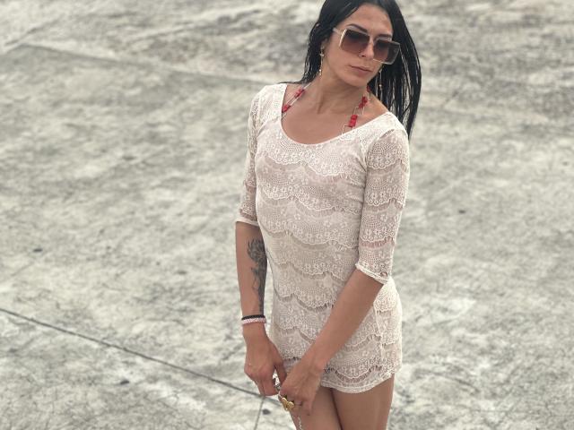 ArielNitzche in a white lace dress, holding a phone, wearing red earrings, brown sunglasses, and a tattoo on her right arm, standing on a gravelly surface.