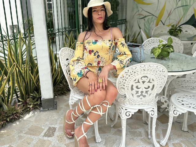 ArielNitzche with dark hair, wearing a yellow floral off-shoulder top, white lace-up sandals, and a sunhat, sitting on a white ornate chair, showing her tits and ass.