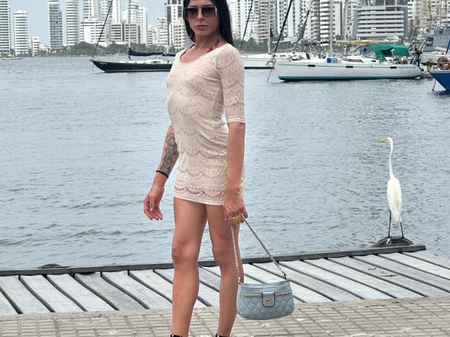ArielNitzche with tan skin, black hair, and tattoos, wearing a lacey beige dress, standing on a dock with a grey purse and a white heron in the background.