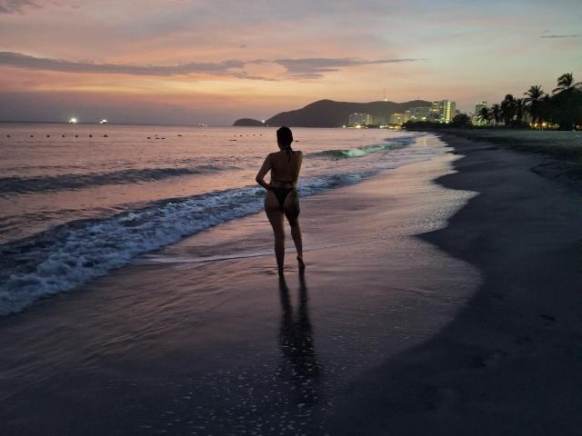 LiaRamonaV with a toned, curvy body, small tits, and a thong, stands in wet sand at sunset, waves gently touching her feet, and a city skyline in the background.