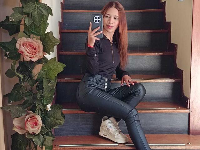 ValerieWoods assise sur les marches, portant un pantalon en cuir noir, un t-shirt noir, des baskets blanches, tenant un téléphone, avec de longs cheveux bruns, des roses et des feuilles vertes sur la gauche, souriante, portant un bracelet rouge..