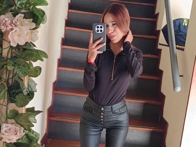 Valerie Woods, con i capelli rossi, una camicia nera con zip e pantaloni di pelle nera, tiene un telefono, è in piedi su delle scale di legno, sorride, mostrando il sedere con i pantaloni abbottonati..