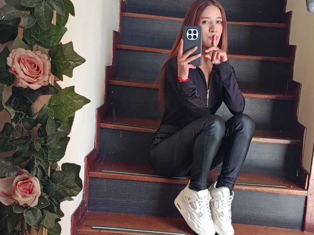  ValerieWoods with long red hair, black jacket, and black leather pants, sitting on wooden stairs, holding a phone, covering her mouth, wearing white sneakers.