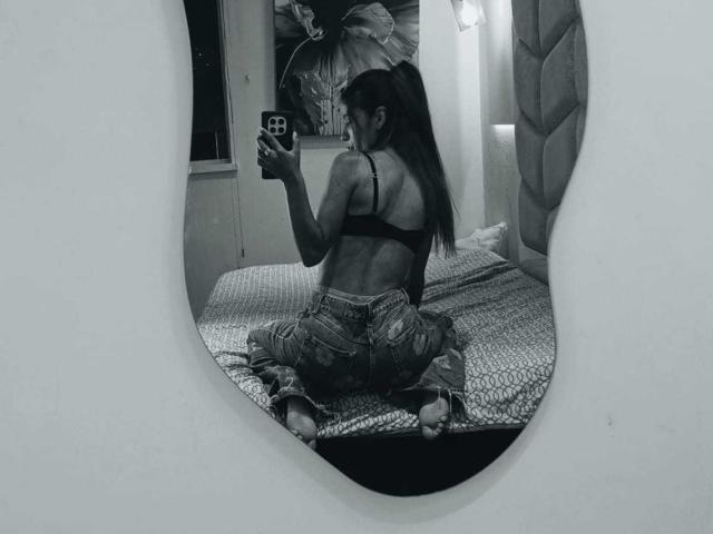 ValerieWoods, back to camera, sitting on bed, wearing black bra, tight jeans, long ponytail, taking selfie, visible ass, mirror reflection, gray-scale, bedroom, bed with patterned blanket.