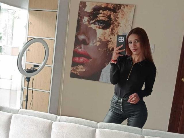 Valerie Woods, con cabello rojo, una camiseta negra con cremallera y pantalones de cuero negro, tomando una selfie frente a un espejo, mostrando su trasero, en una habitación con una luz de aro y un fondo para retratos..