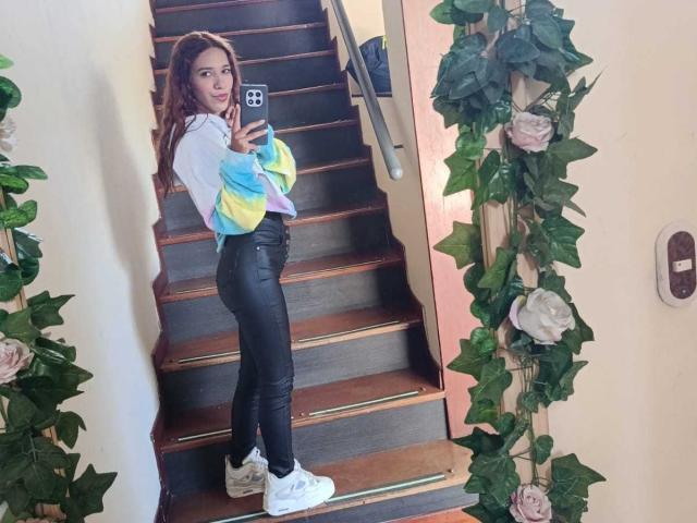 Valerie Woods, con cabello largo y castaño, sudadera de algodón con estampado de arcoíris, pantalones de cuero negro, zapatillas blancas, de pie en las escaleras, tomando una foto de sí misma en un espejo, enredaderas de hojas verdes, rosas rosadas en las paredes..