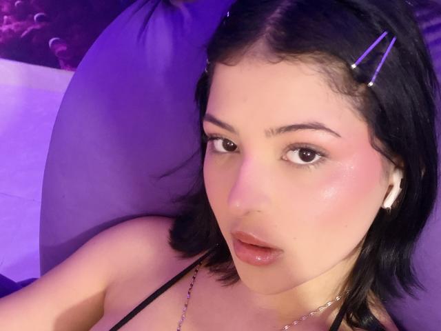 RheaVolk with shiny pink lips, black hair with purple hairpins, wearing a black strap, looking at the camera, pink blush on cheeks, white earbuds, purple pillow.