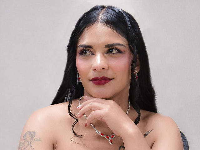 Mia Bellucci a de longs cheveux noirs, du rouge à lèvres rouge et des boucles d'oreilles rouges, sa main est près de son visage, révélant un petit tatouage sur son épaule, et elle porte un collier avec des pierres rouges..