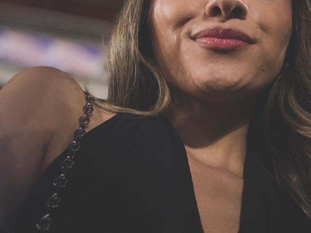 Il viso di AdharaCooperr, con un sorriso appena accennato e rossetto rosa, indossa un abito nero senza maniche con dettagli simili a perline sulla spallina. I suoi capelli castani ondulati le ricadono sui lati..