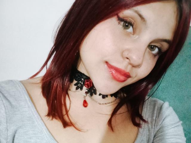 SophieHill with red lipstick, black lace choker, and red rose, wearing a grey top, has a cute nose ring, and red shoulder-length hair, smiling at the camera.