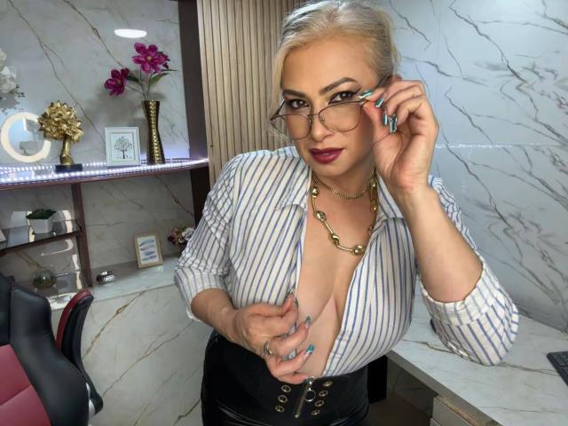 MisseScarleth with blonde hair, glasses, and a deep cleavage in a striped shirt, holding a phone to her ear, wearing a black corset and gold necklace, smiling.