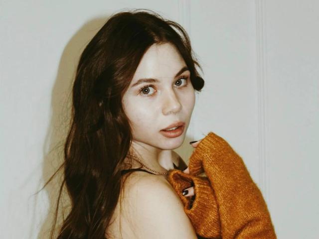 Azzie Stelman, con lunghi capelli castani, occhi verdi e un leggero sorriso, indossa un maglione arancione senape, tenendolo sopra il seno, e guarda indietro verso la fotocamera..