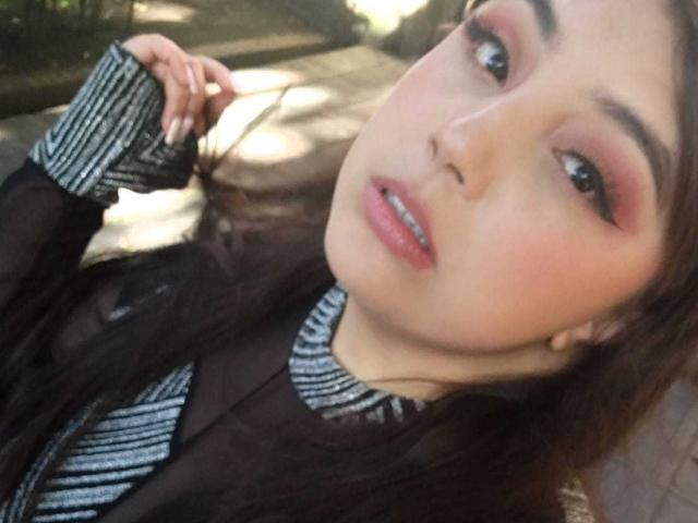 RoxannDiaz with pink eyeshadow, black striped shirt, and glossy pink lips, her hand in her brown hair, looking at the camera with a slight, sexy smile.