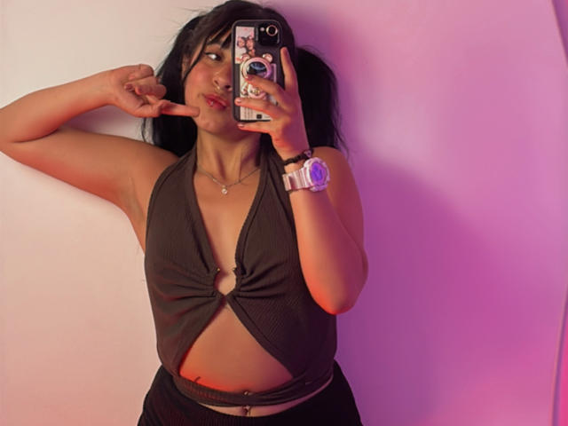 MiaPresscot com maria-chiquinhas, blusa de alça preta que mostra seus seios, e um anel, calças pretas, luz rosa-roxo, dedo perto dos lábios, segurando um celular com capa de anel, usando um relógio..