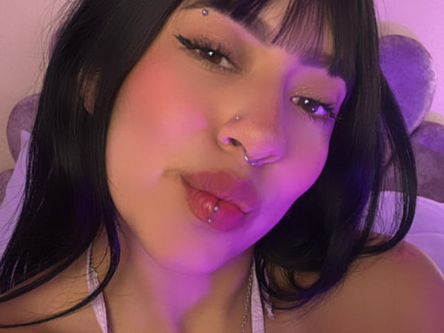 MiaPresscot hat schwarzes Haar, rosigen Lippenstift und einen Nasenring mit einer Septum-Piercing. Sie zieht die Lippen zusammen, trägt einen weißen Top mit Halstiefe und befindet sich unter violettem Licht..