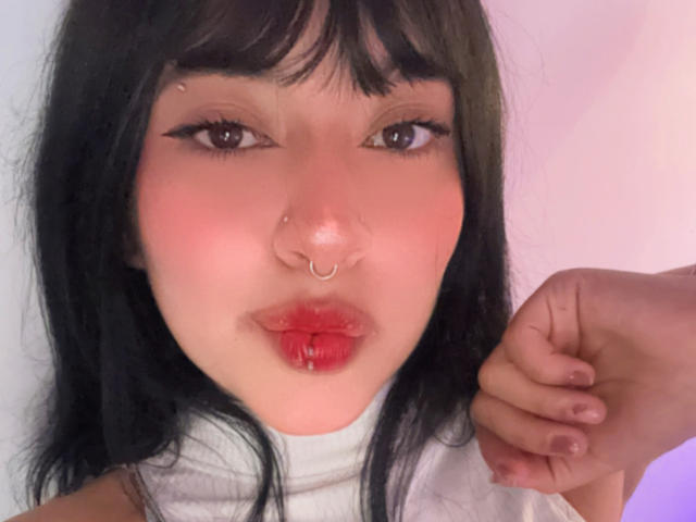 MiaPresscot a les cheveux noirs, les yeux marron, du rouge à lèvres et un piercing au nez. Elle plisse les lèvres avec une main près de son visage, montrant un blush rose sur ses joues..